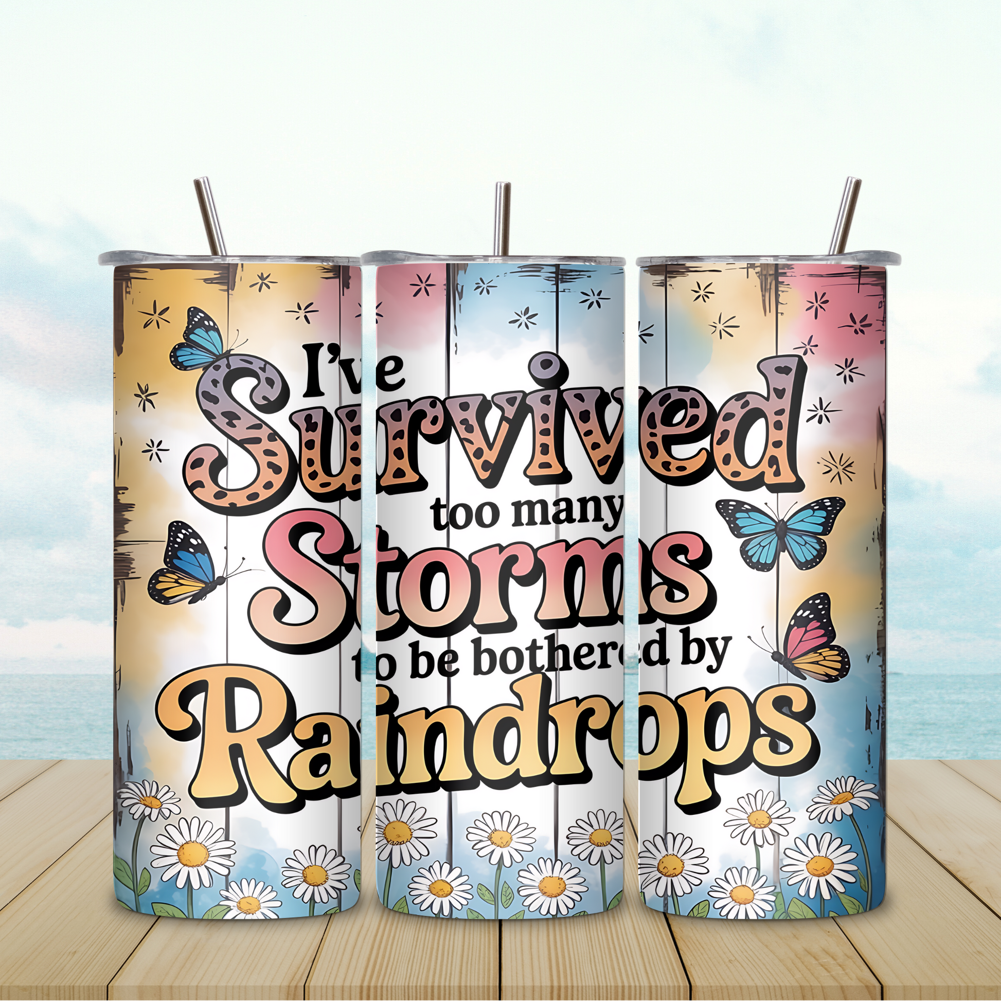 Three colorful tumblers with motivational text and butterfly designs on a wooden surface with a scenic background.